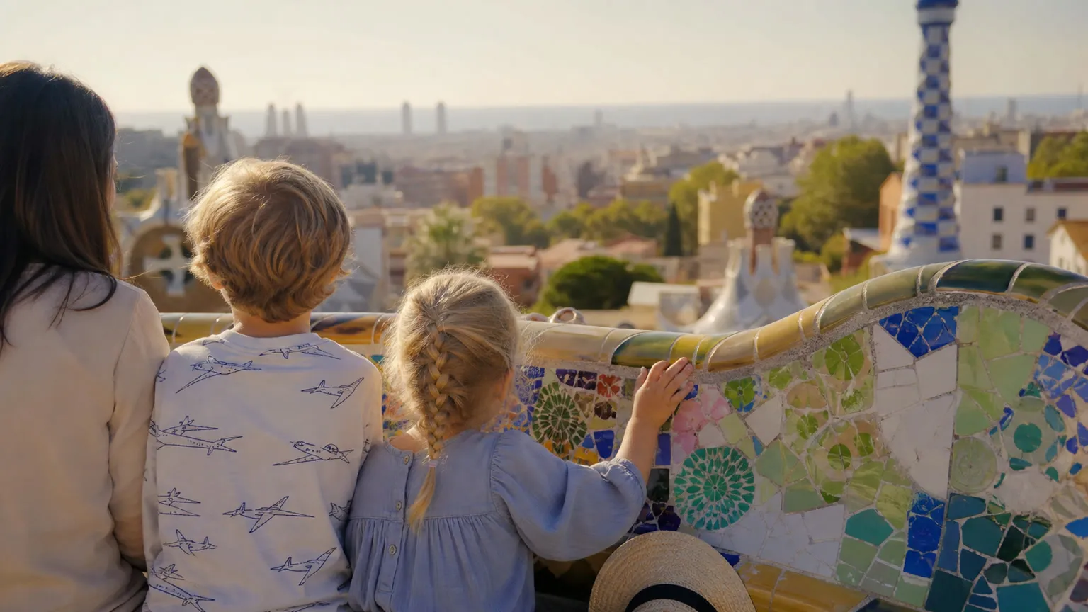 Børn set bagfra ved Park Güells mosaikbænk med Barcelonas skyline i morgensol