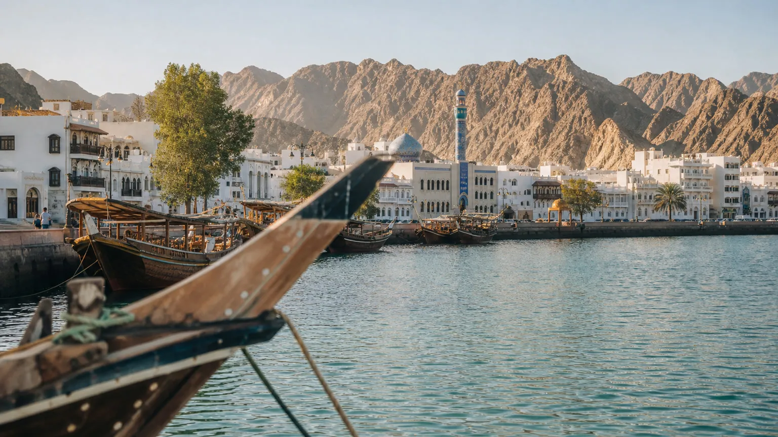 Dhow-både ved Muttrah Corniche i Muscat med turkis bugt, hvide bygninger og Al Hajar-bjergene i baggrunden