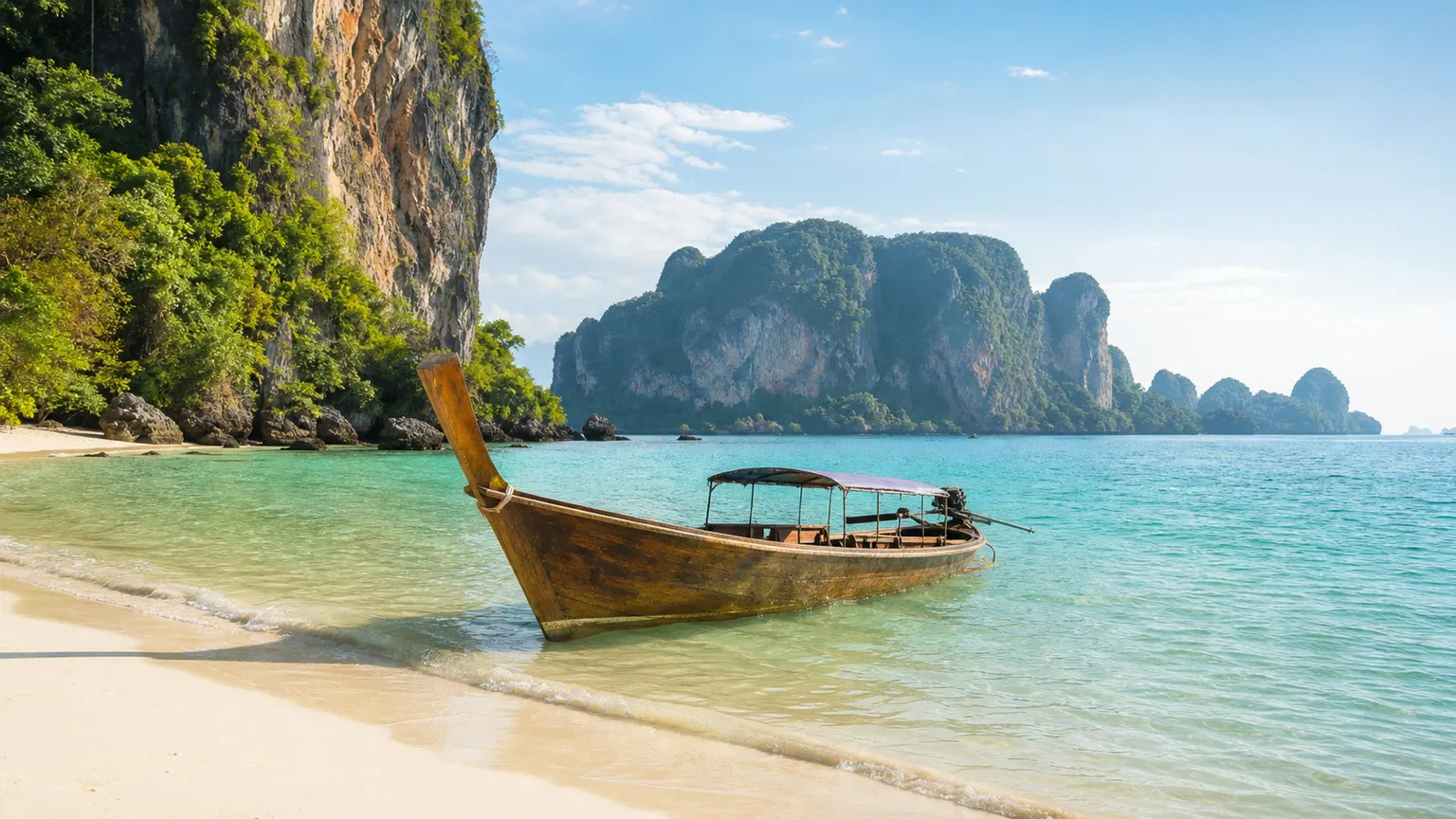 Longtailbåd i turkisblåt vand ved hvid sandstrand og kalkstensklipper i Thailand